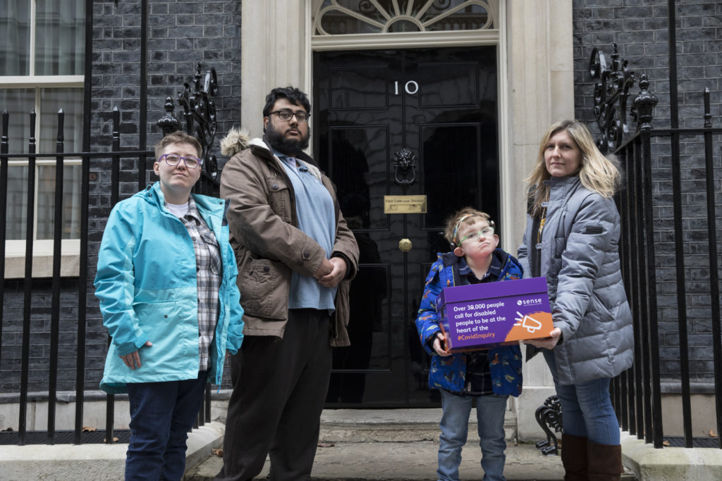 Landscape shot of three adults and one child standing in front of Number 10, Parliament. Their expressions are serious and the boy holds a purple and orange box with writing on the side reading 'Over 39,000 people call for disabled people to be at the heart of the #CovidInquiry'