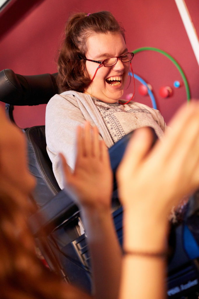 A young woman wearing glasses laughing, with hands clapping in the foreground.