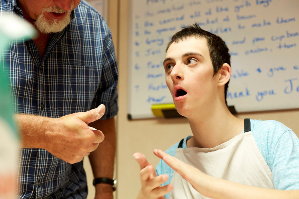 A young man sits in front of a whiteboard, while his support worker gives him a thumbs-up.