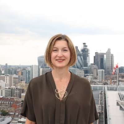 A woman with cropped blonde hair and a smart top and necklace is pictured with the London skyline in the background