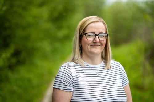 A woman in a stripy top smiles at the camera. There are bushes and grass behind her.