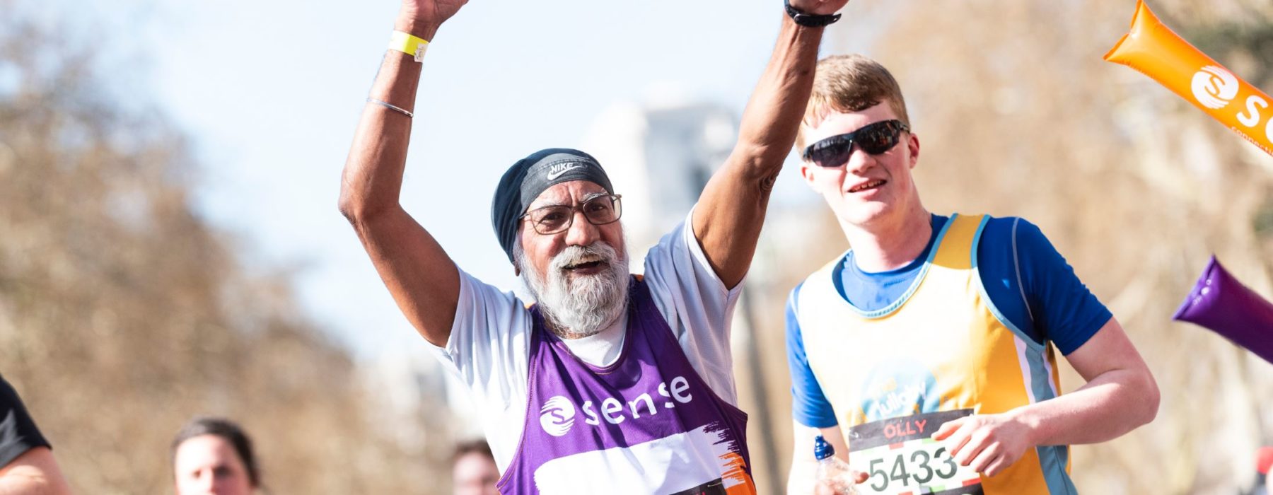 A older man in a Sense running vest holds his hands as he runs in a marathon