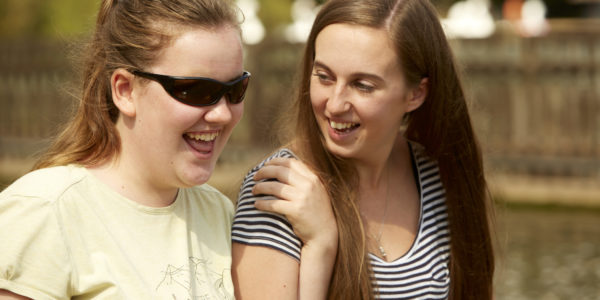 Two women are smiling and walking, one is wearing dark sunglasses