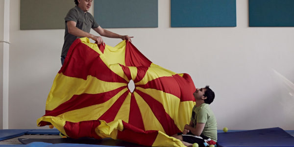 A man is holding a red and yellow parachute next to a young man who's sitting on the floor cross legged