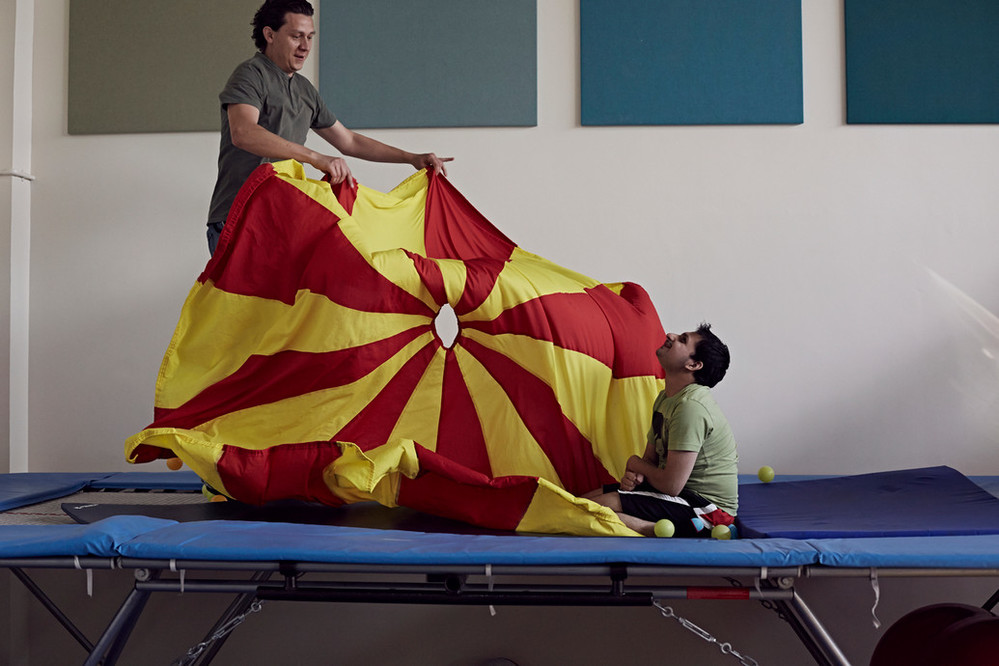 A man is holding a red and yellow parachute next to a young man who's sitting on the floor cross legged