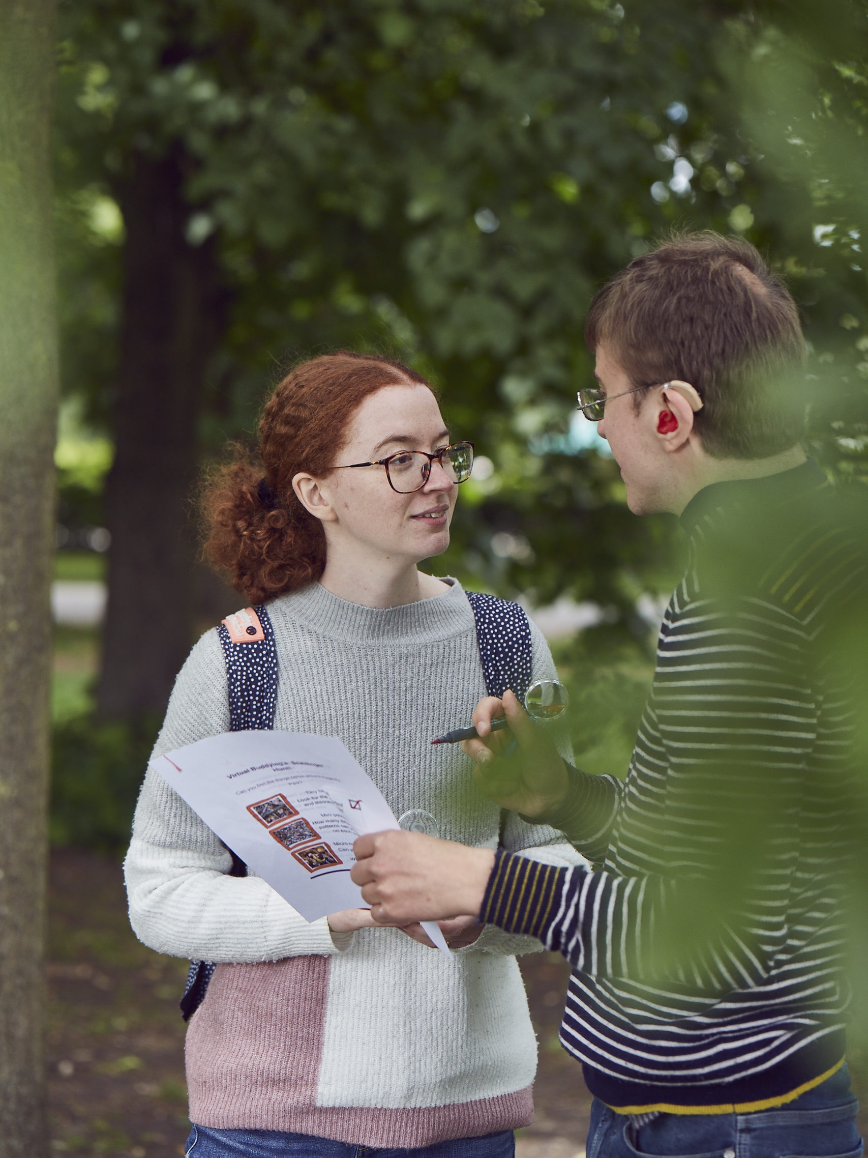 A red-haired woman and a brown-haired man are talking next to some trees
