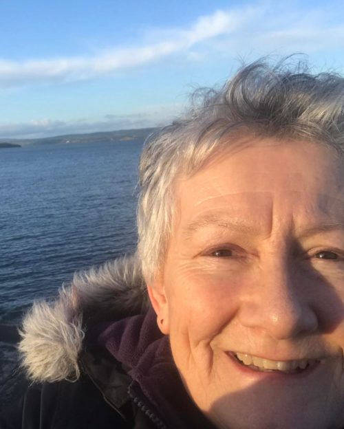A woman with short grey hair has a coat on and is smiling. She's standing in front of the sea.
