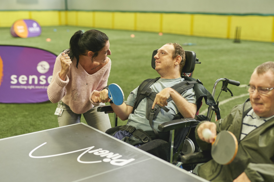 A white male in a wheelchair holds a table tennis bat whilst a white woman with brown hair supports him