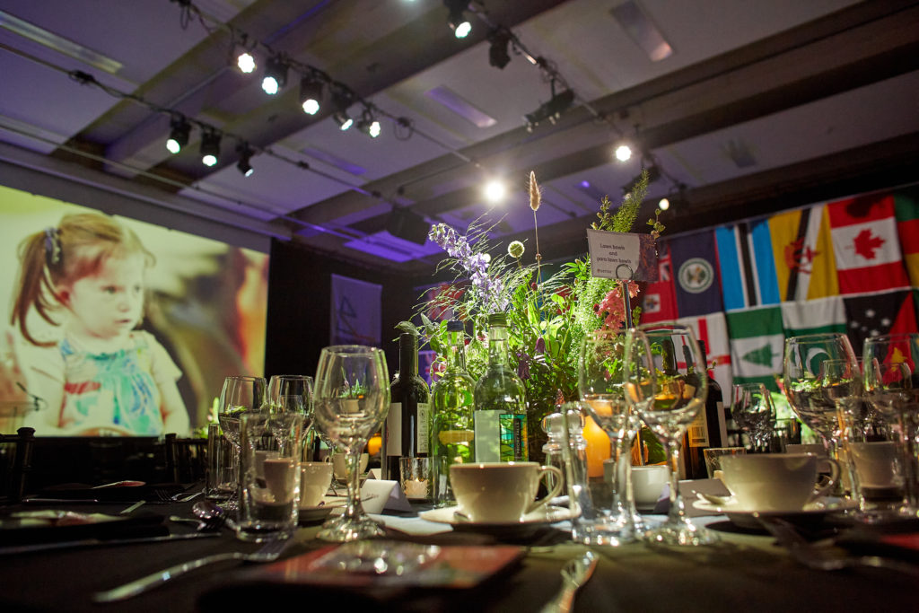 A large room in TouchBase Pears is decorated for the gala dinner. Tables are covered in a black table clothe and laid with cutlery, glasses, candles and wild flower centre pieces. Behind the tables we can see flags handing from the ceiling and a large screen with a Sense video projected onto it.  