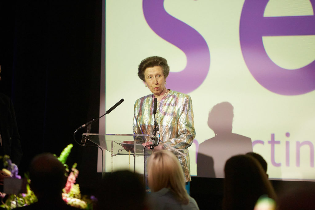 HRH The Princess Royal stands on a stage giving a speech. Her hair is dark brown and styled up. She is wearing a metallic patterned jacket and behind her we see the Sense logo projected onto a large screen.  
