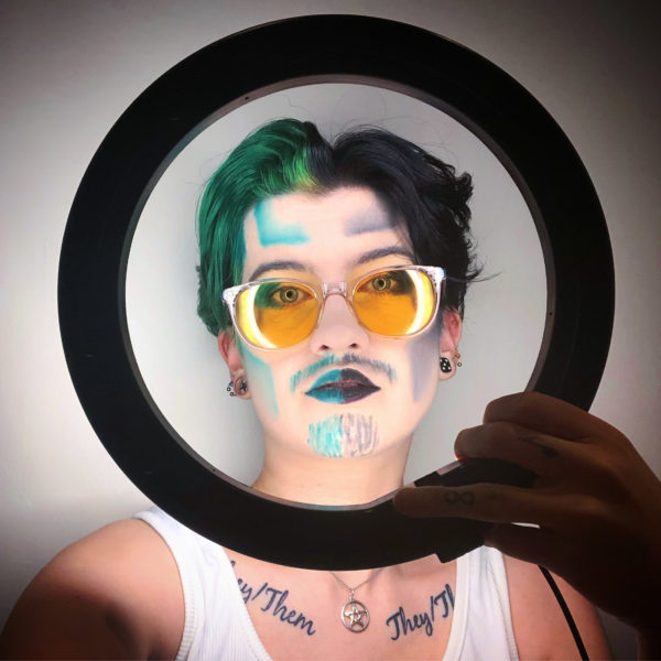 Papa Smear is taking a selfie while holding a ring light up to his face. His face is half and half black and green to match his split dyed hair, with the left half having green contour, moustache, beard, lips, and eyeshadow, and the right having black contour, moustache, beard, lips, and eyeshadow. He is wearing his glasses with orange tinted lenses. You can see his they/them collar bone under the ring light, and he’s wearing a white vest.