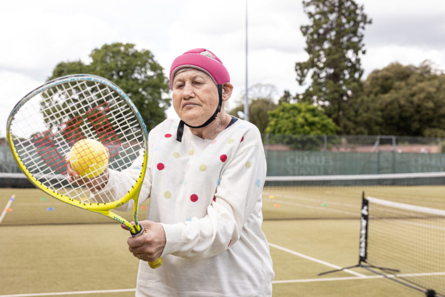 A woman stands on a lawn holding a tennis racket and a ball