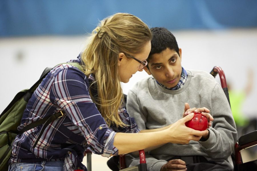 Young boy in a wheelchair wearing grey jumper holds a boccia ball, assisted by support worker