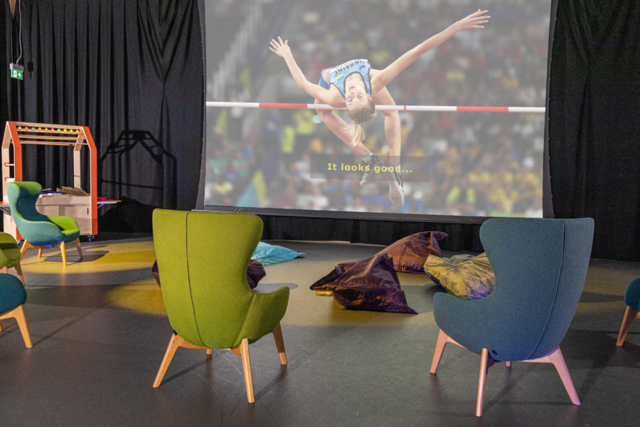 Two large comfy chairs are facing a big projector screen, one is green and one is blue. On the screen is a Ukrainian athlete doing the high jump. Between the chairs and the screen are lots of soft bean bags on the floor of the room.
