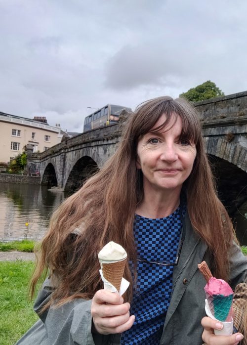 Tracy, a white woman with long brown hair, is standing holding a vanilla ice cream. Someone else, who we can't see, is holding a strawberry ice creamwith a chocolate flake. She's standing in front of a river with a big old bridge over it. The weather is grey, but Tracy looks like she's having fun.