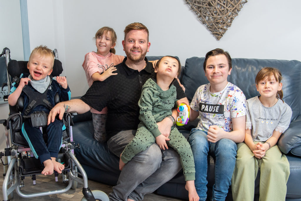 A man sits on a sofa with his young children. One son is on his lap, his daughter holds his shoulders, next to him on the sofa are another son and daughter. One son in a wheelchair sits next to the sofa holding dad's hand.