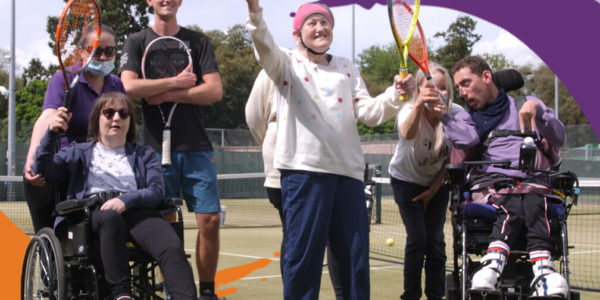An older woman stands holding a racket and tennis ball, with her arm raise. Two people in wheelchairs and two standing coaches stand either side of her
