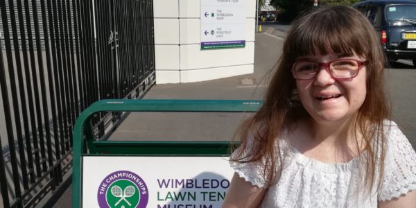 Sian, a young woman with long brown hair, stands smiling in front of a Wimbledon Lawn Tennis Museum sign.