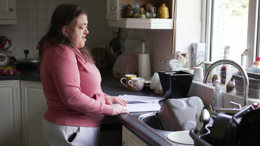 A woman stands at her kitchen counter, reading a bill in braille, with the kitchen sink in the foreground.