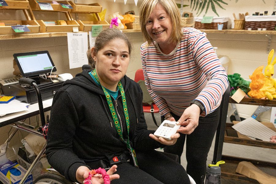 Two women looking to camera, one is in a wheelchair, one is standing. They are in a shop. The woman in the wheelchair is holding a card machine in her hand