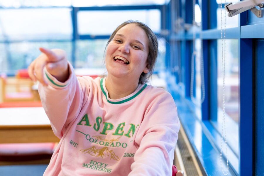 A young woman wearing a pink jumper smiles and points to the camera.