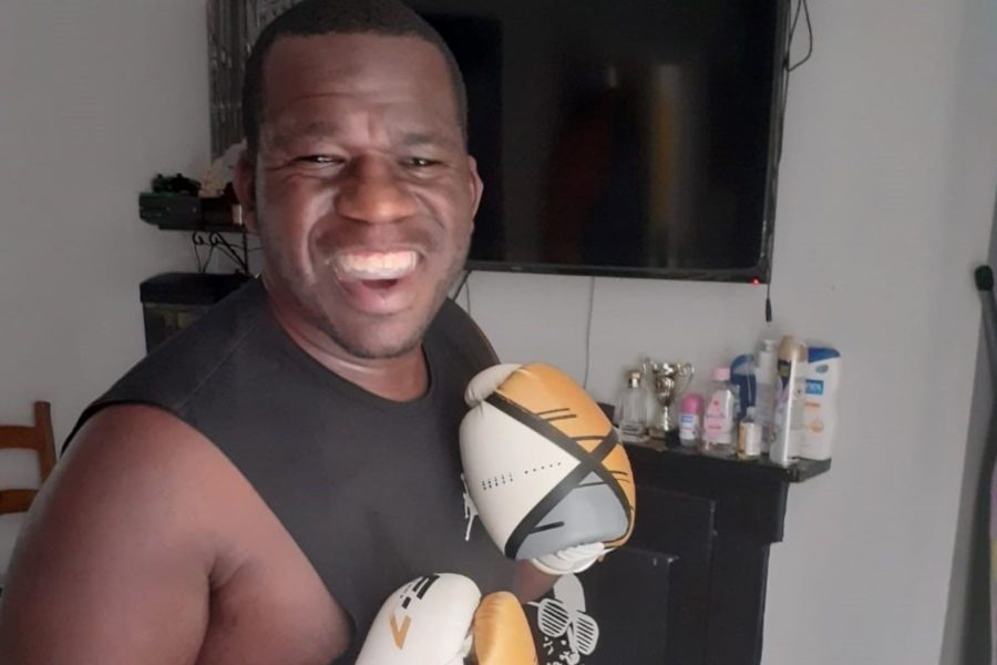 A man wearing a black vest and white and gold boxing gloves, smiles widely.