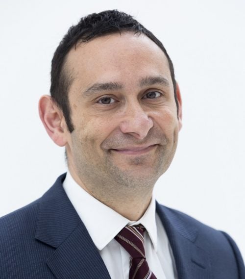 A white man with dark hair wears a suit and smiles at the camera