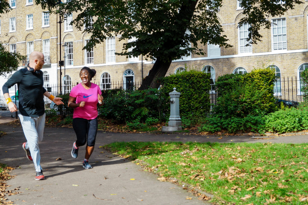 James and Nike, a white man and a black woman dressed in athletic clothes, running through a park and smiling at each other.
