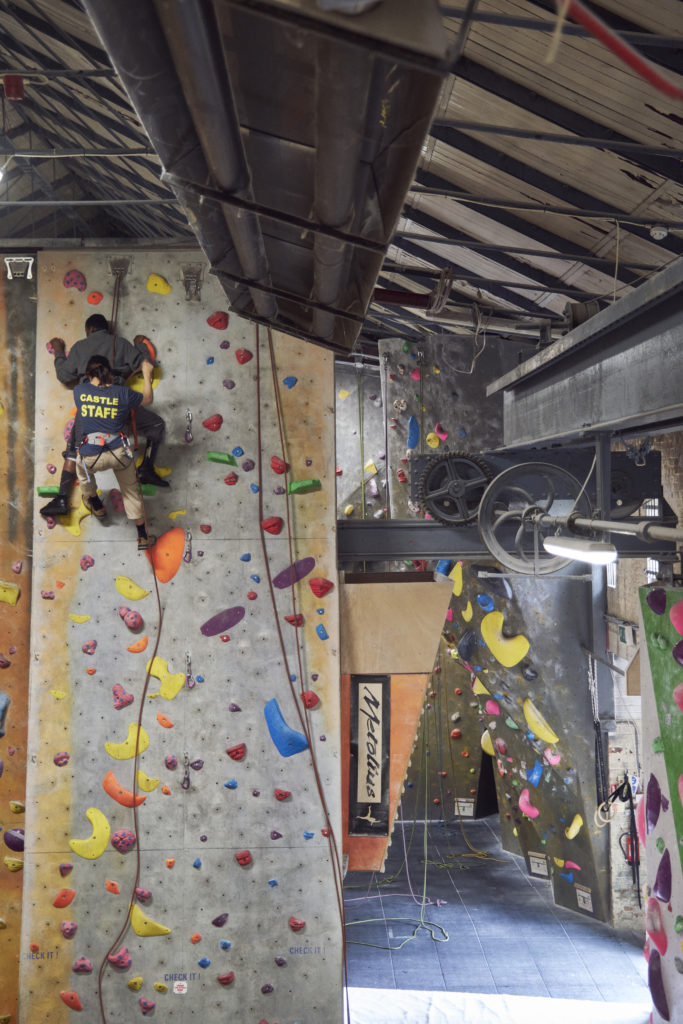 A tall indoor rock climbing wall in a large, warehouse like room. Its covered in multi coloured raised surfaces of different shapes and sizes to climb on.