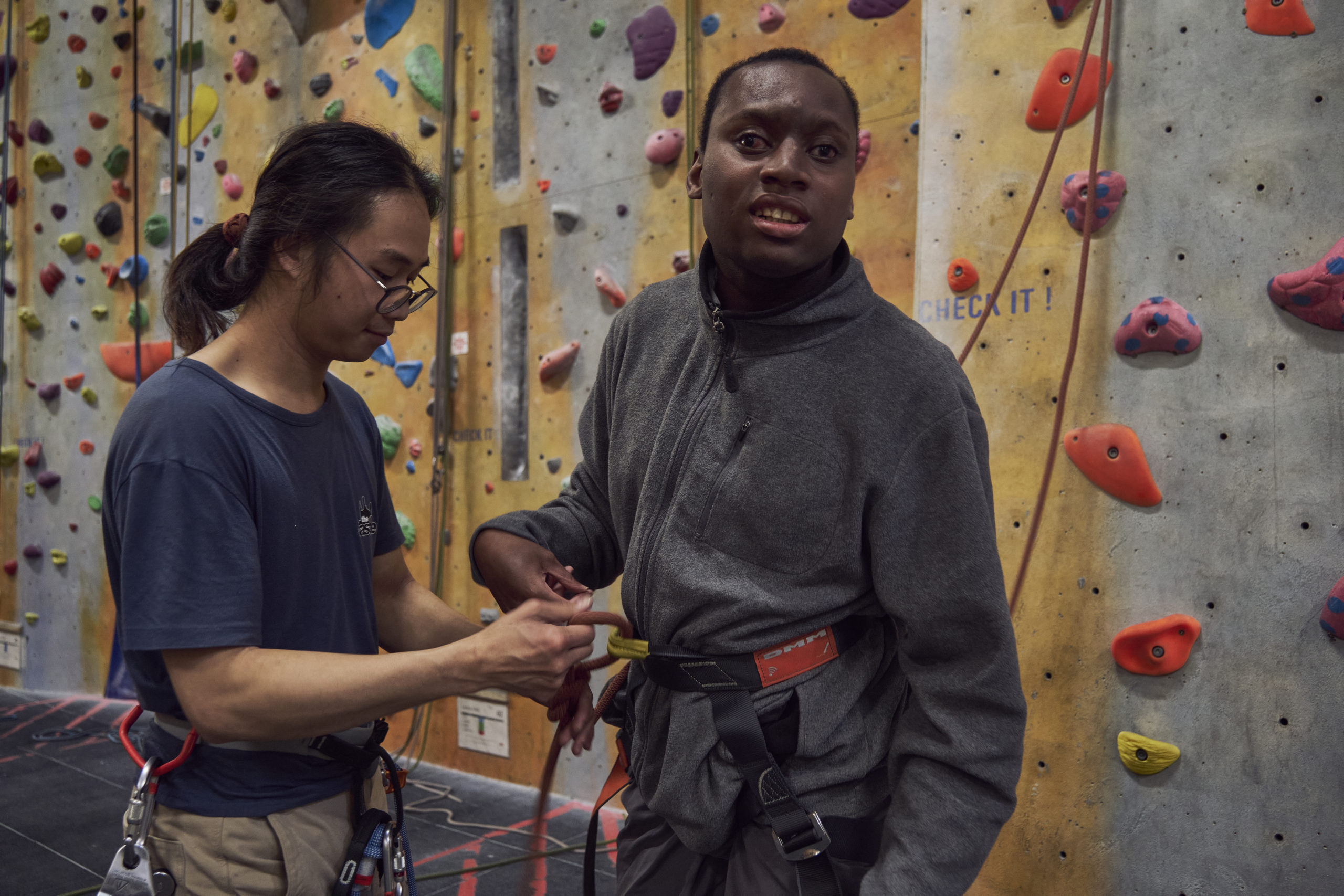 A young man stands in front of a climbing wall with brightly coloured grips. Another man helps him to attach his harness.