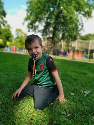 A boy in a football top sits on grass with his dummy in