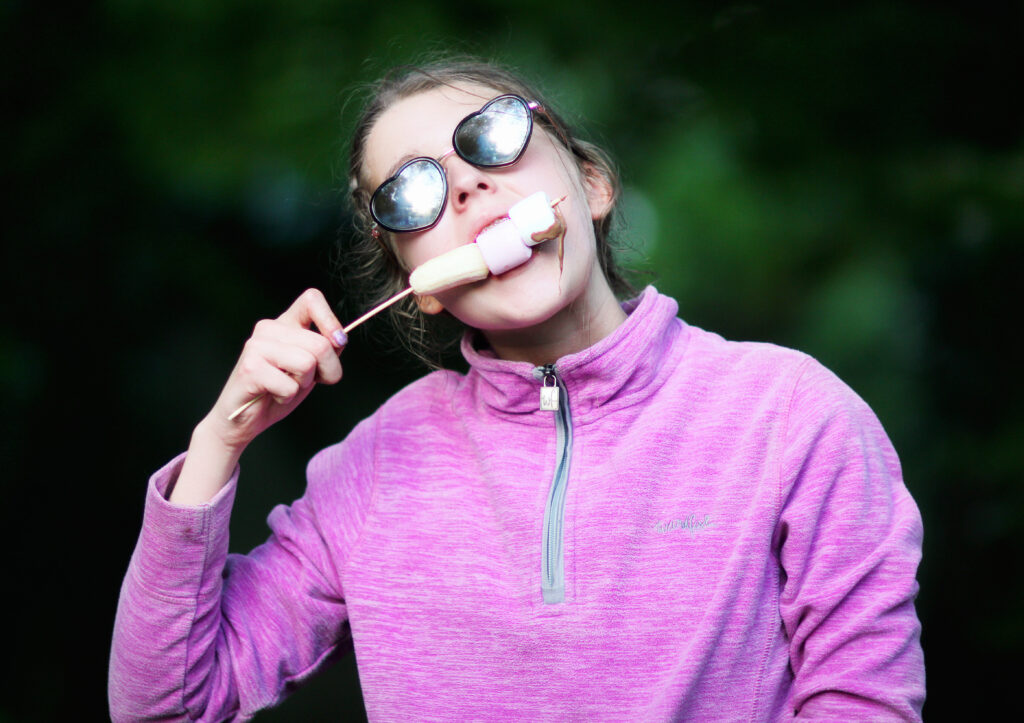 A young girl in a bright pink top wearing sunglasses is eating marshmallows off a stick.