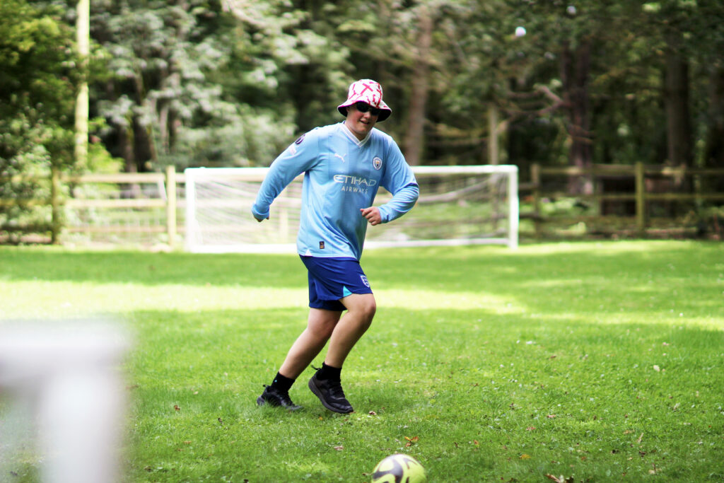 A young man wearing a blue football kit and a bucket hat is running towards a football in a field. Behind him are white goalposts and trees. 
