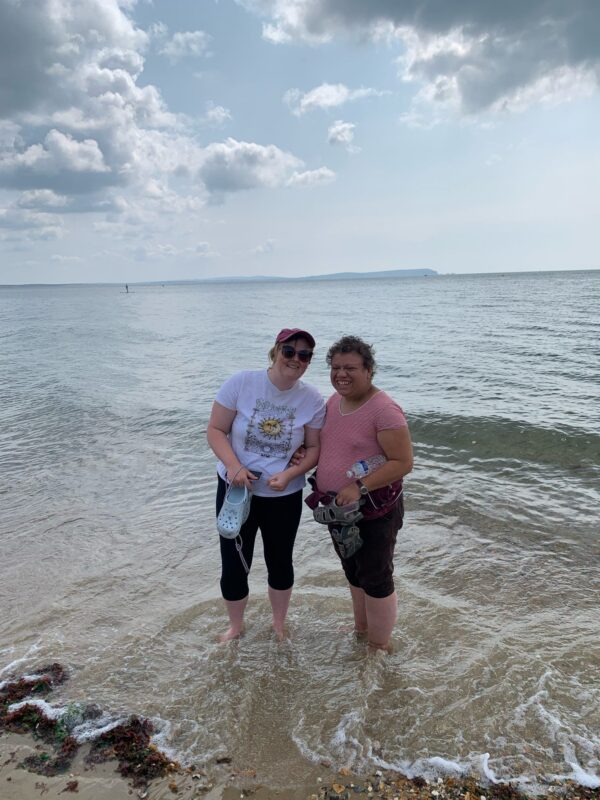 Two women are standing in the sea, they have their trousers rolled up and the waves around their ankles. 