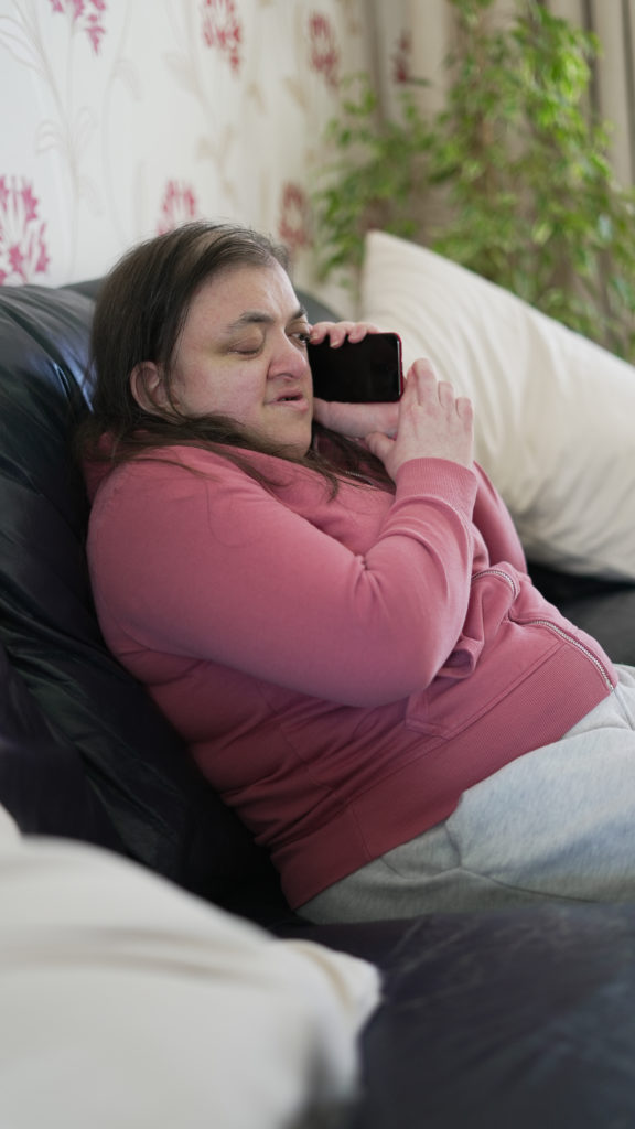 A woman in a hoodie sits on the sofa with her phone held to her ear.