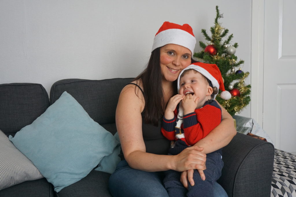 A little boy and his mum, both wearing Santa hats, sit smiling in front of a Christmas tree.