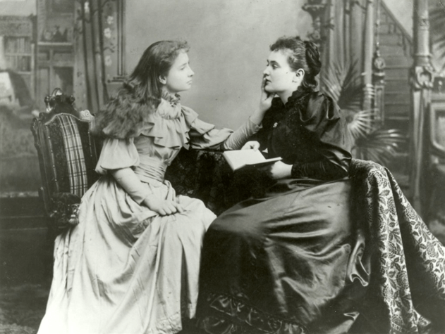 A black and white image of Helen Keller, touching the face of a companion who is reading a book.