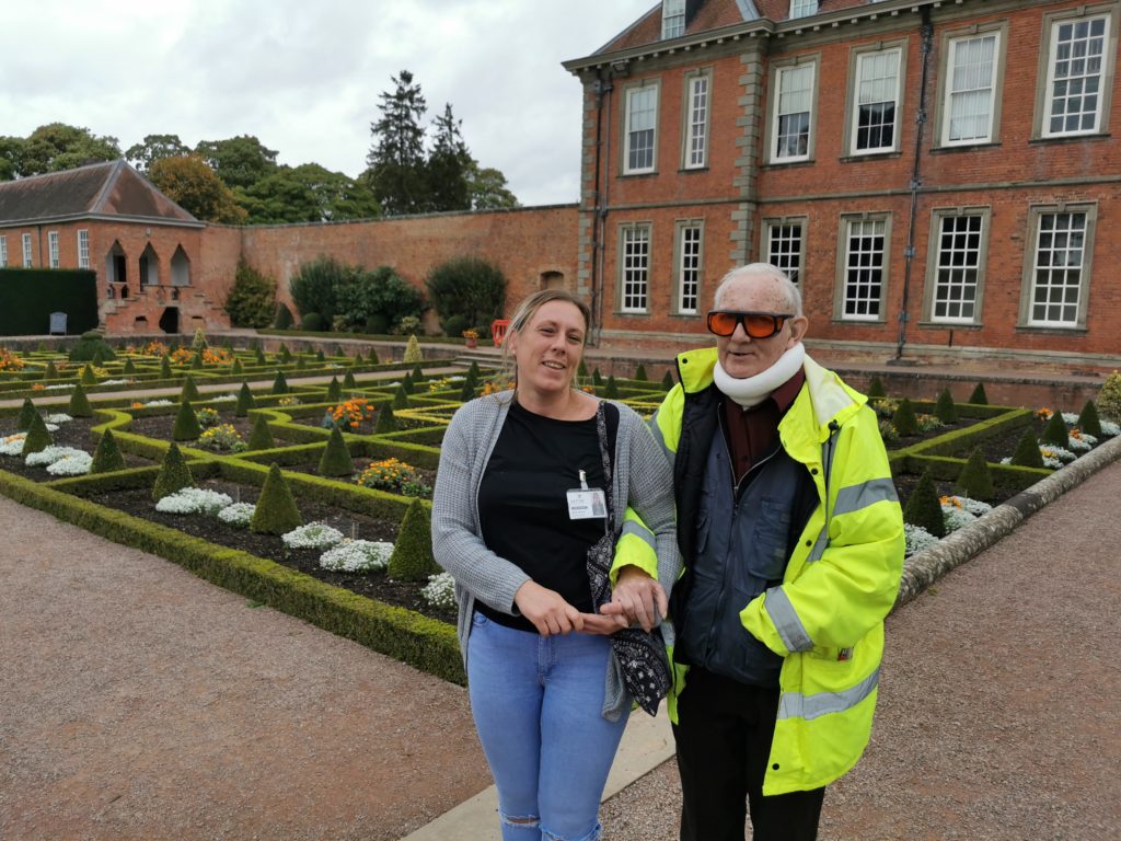 David, a man wearing a hi vis jacket and a neck brace, walks arm-in-arm with a woman through a garden.
