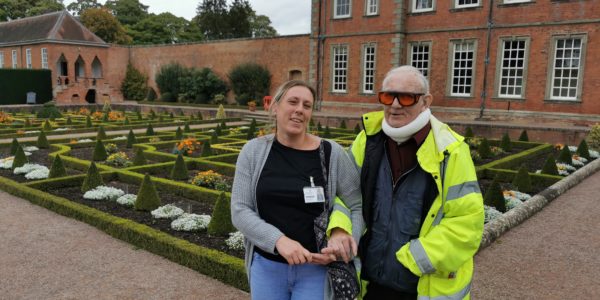 David, a man wearing a hi vis jacket and a neck brace, walks arm-in-arm with a woman through a garden.