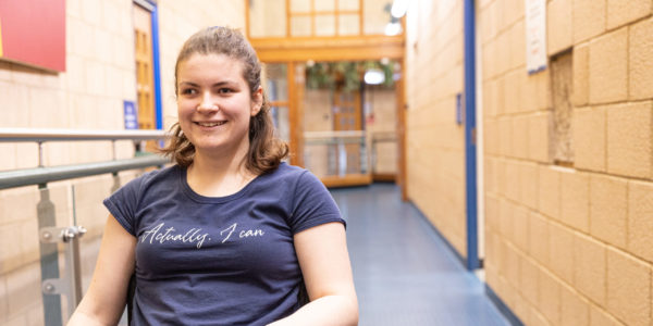 A young woman in a wheelchair wearing a blue tshirt smiles to camera in an indoor space.