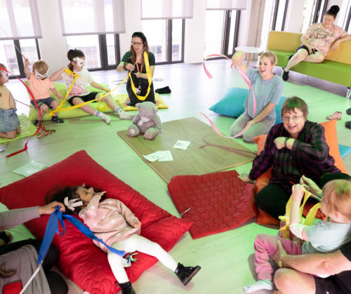 A group of children and adults sit on the floor or on bean bags and play with colourful toys and ribbons