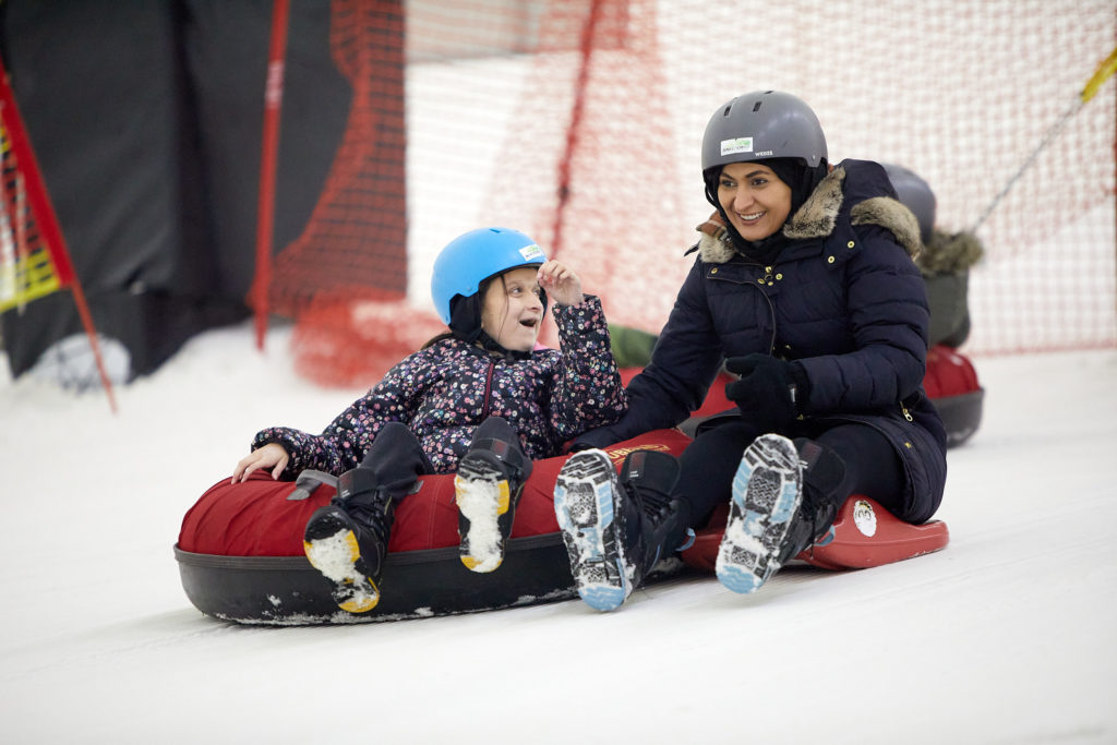 A young person and support worker both smile while sledging down a ski slope.