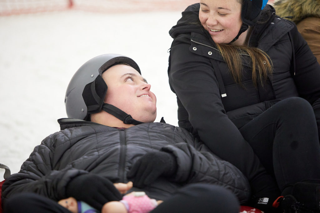 Two young people wearing helmets and ski clothes smile at each other.
