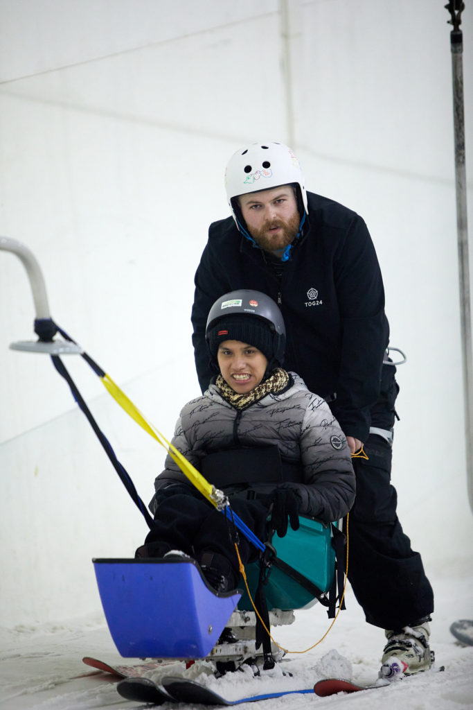 A young person in a sit ski is being hoisted uphill, with a ski instructor behind him.