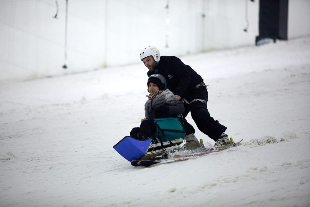 A young person rides a sit ski down a slope, supported by a ski instructor.