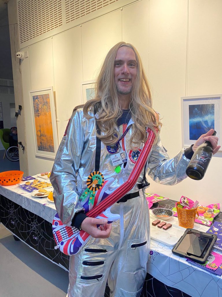 A man in a fancy dress costume wearing a long blonde wig and a silver space suit, holding an inflatable guitar. 