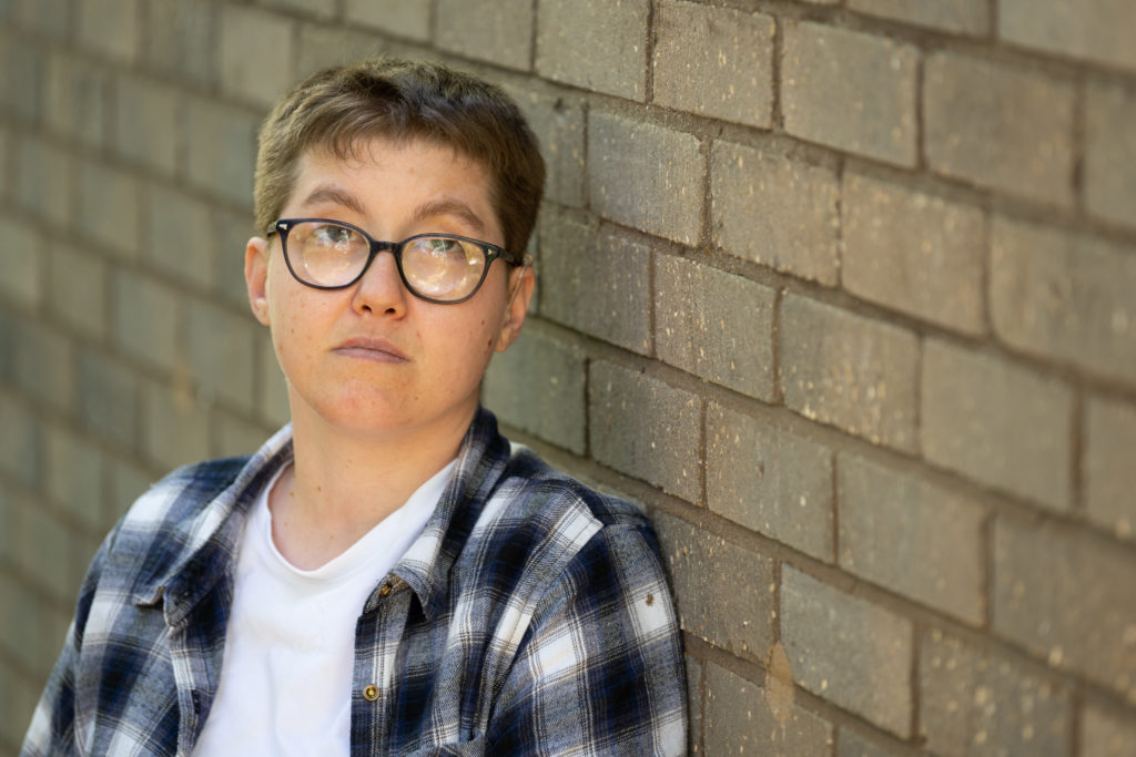 A close-up of Emma, a white woman with short cropped hair and glasses, standing in front of a brick wall.