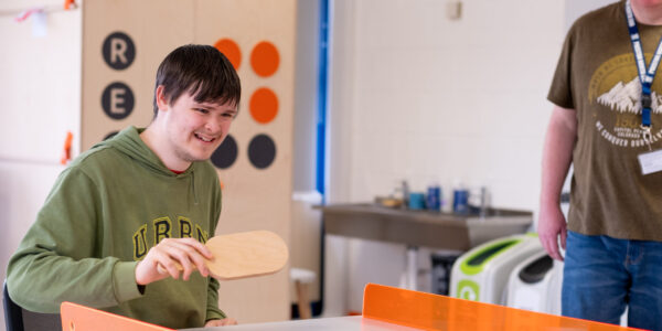 A young man wearing a green hoodie is playing polybat, a game involving a wooden bat, a ball and a wooden table with orange sides.