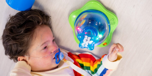 A young boy with dark brown hair wearing a sweatshirt with a rainbow on lies down whilst playing with a sensory toy