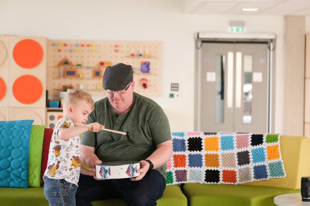 Thomas, a small boy with blonde hair, and his dad playing with a drum in Sense's children's room at TouchBase Pears in Birmingham. 